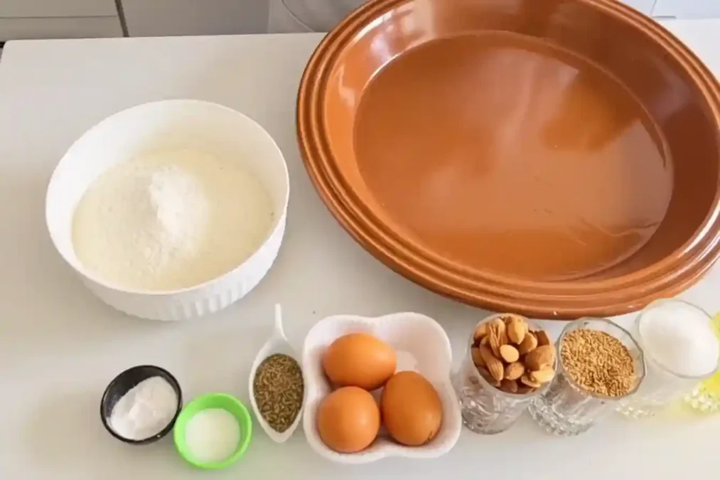 Ingrédients du fekkas marocain disposés sur une table avec farine, œufs, amandes, graines et sucre.